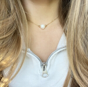 Necklace with a pearl on the neck of a woman with blonde hair and wearing a white zip up jacket
