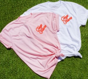 Two shirts on top of grass, one light pink, the other white with the text "Tengo Chisme" in the left pocket area in red